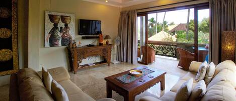 Amplia sala de estar con un sofá grande, una mesa de centro y un televisor. Balcón con vista a palmeras y casas.