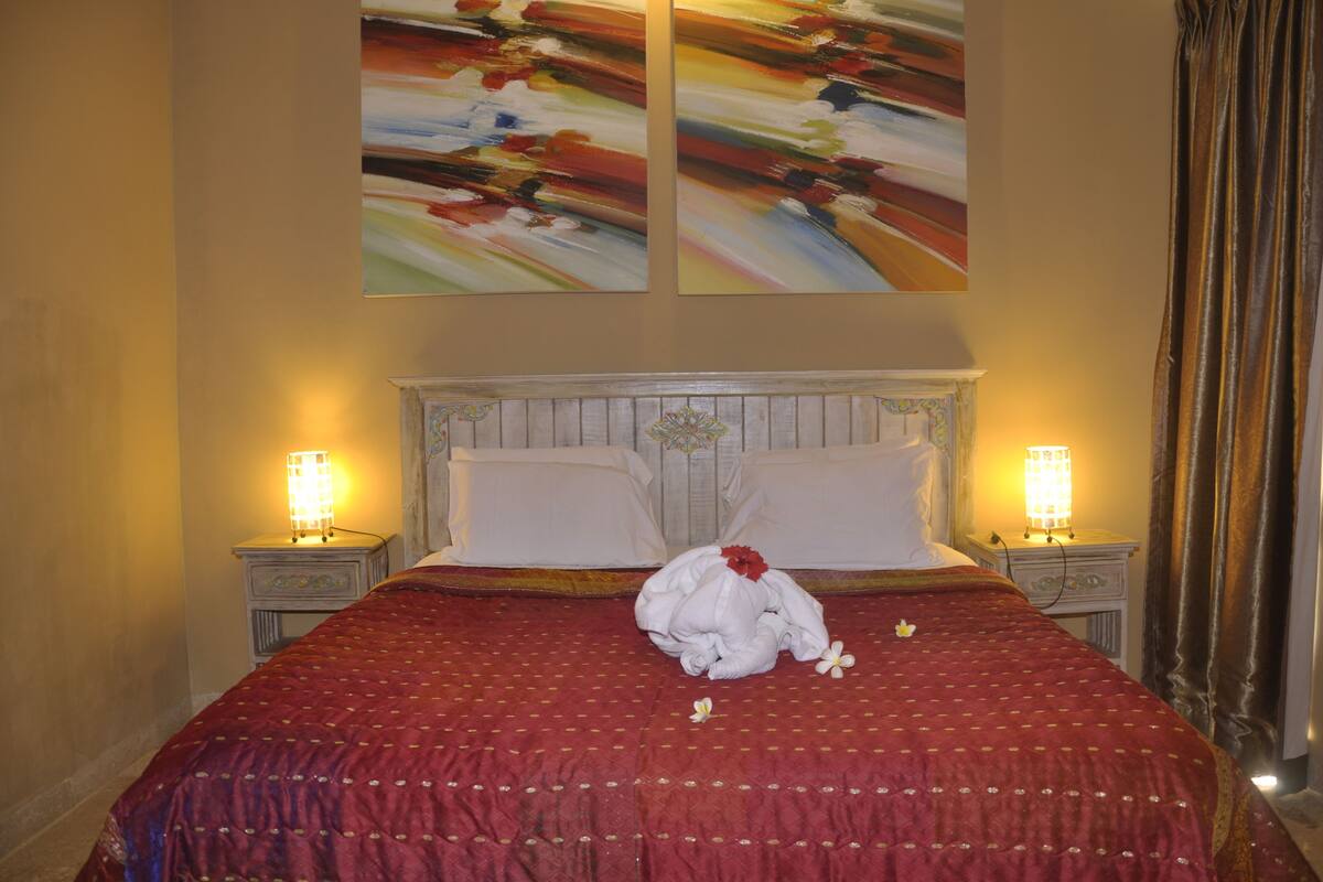 Een net opgemaakt bed met een rode sprei, twee witte kussens en een decoratief object op het bed. Aan weerszijden van het bed staan twee nachtlampjes.