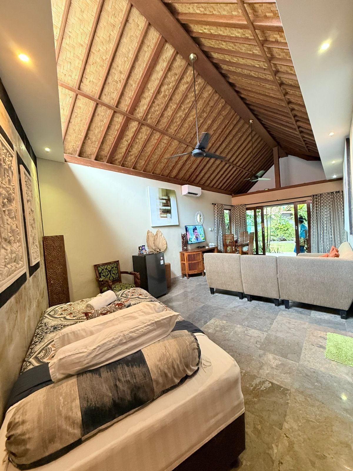 A spacious room with a high ceiling, a bed with patterned bedding, a sofa set, a television, and a ceiling fan.