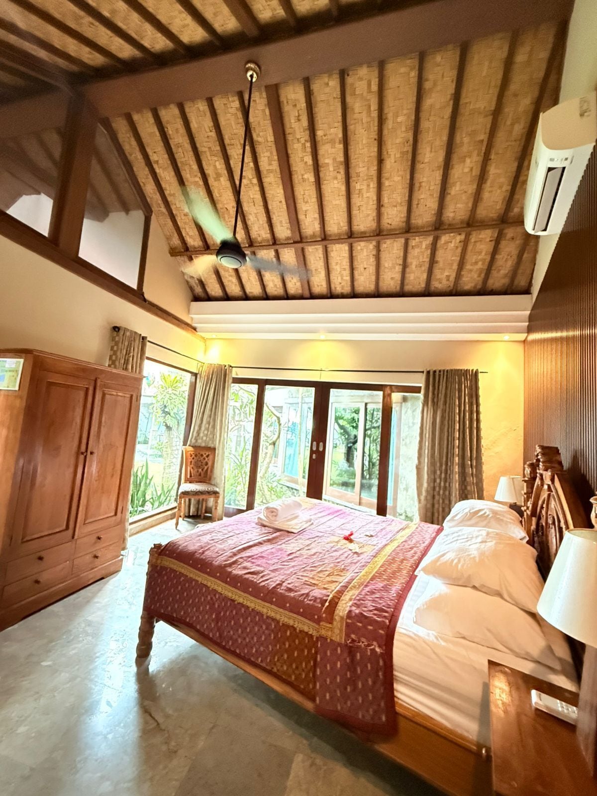 A bedroom with a large bed, wooden ceiling beams, a wardrobe, and a window with a view of greenery.