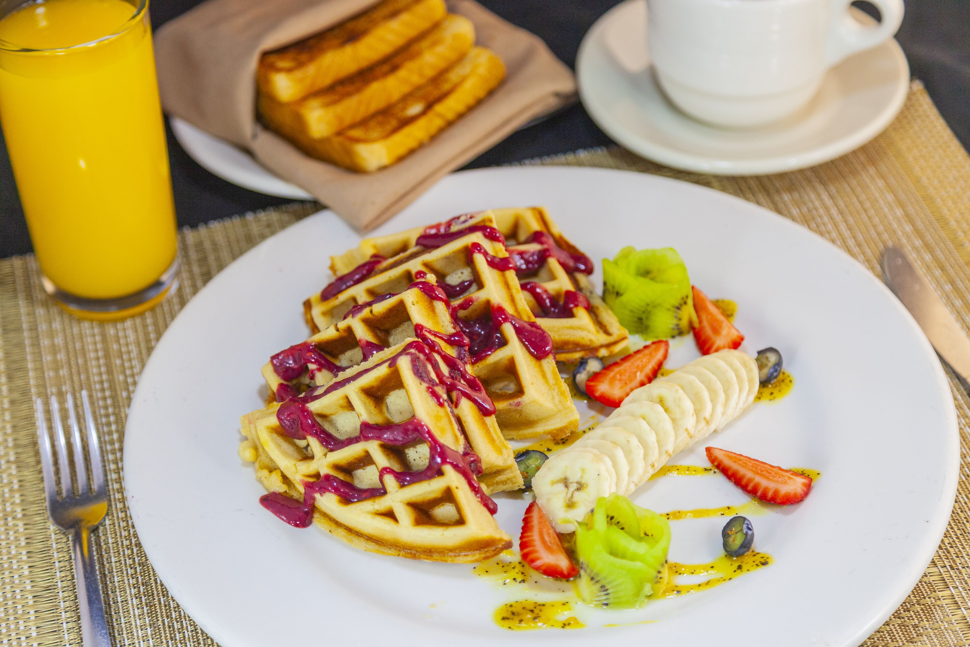 A breakfast plate with waffles, strawberries, kiwi, and banana slices, accompanied by a glass of orange juice and a cup of coffee.