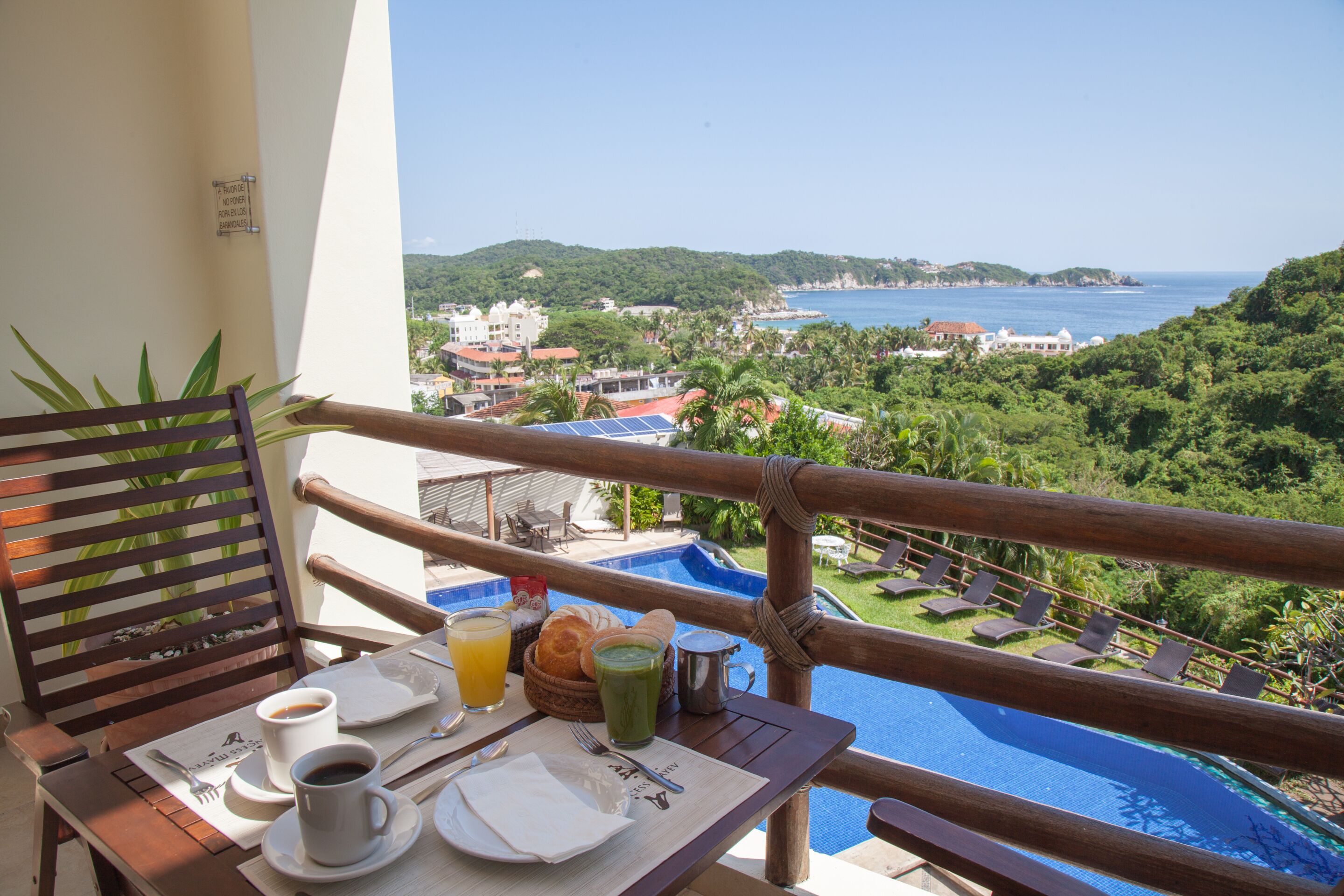 A balcony with a table set for breakfast, overlooking a pool and a coastal view.