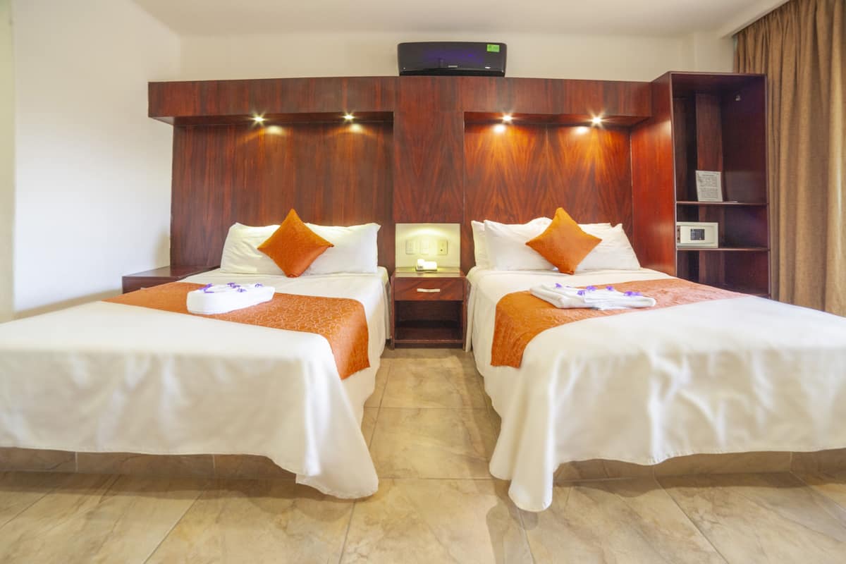 A hotel room with two beds, each with white bedding and orange accents, a wooden headboard, a nightstand with a lamp, and a built-in shelf.