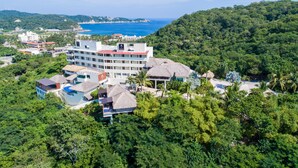 Un complejo turístico con varias edificaciones, incluyendo un hotel grande con techo rojo, rodeado de abundante vegetación y vista al océano.