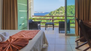Habitación de hotel con cama, mesa y sillas, y un balcón con vistas al mar y vegetación.