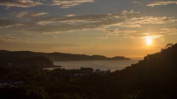 A coastal landscape with a sunset over water, hills in the background, and buildings in the foreground.