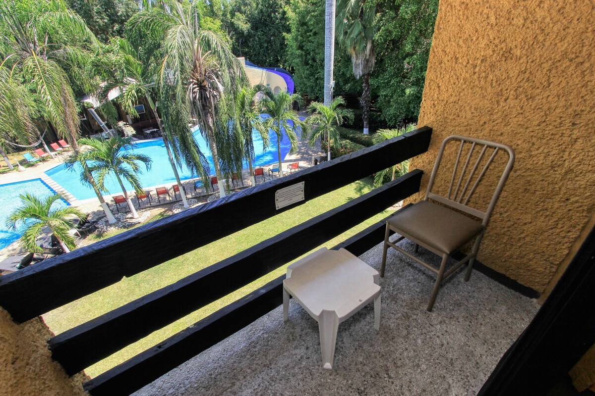 Un balcón con vistas a una piscina, palmeras y una silla.