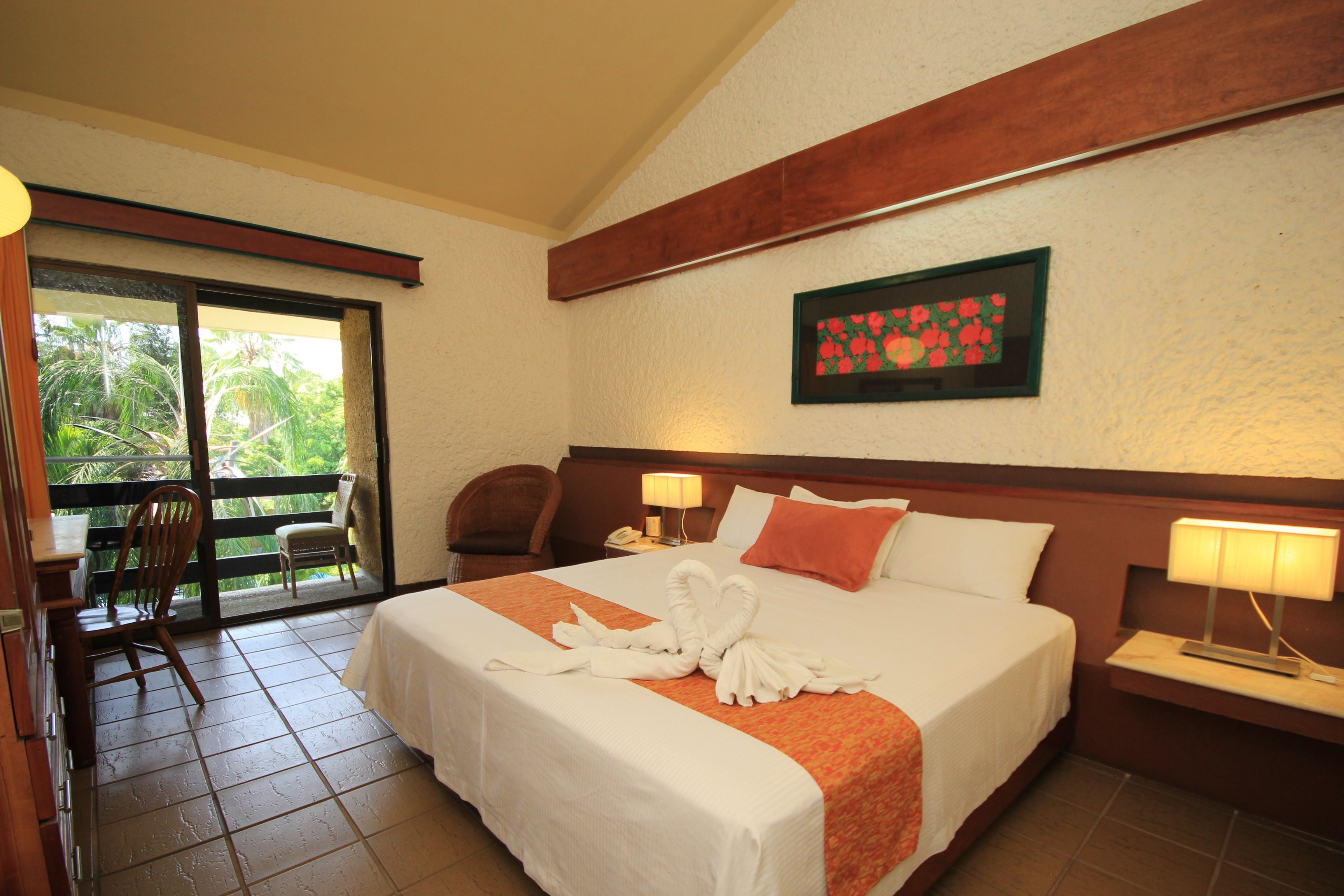 A hotel room with a bed, a chair, a desk, and a balcony with a view of greenery.