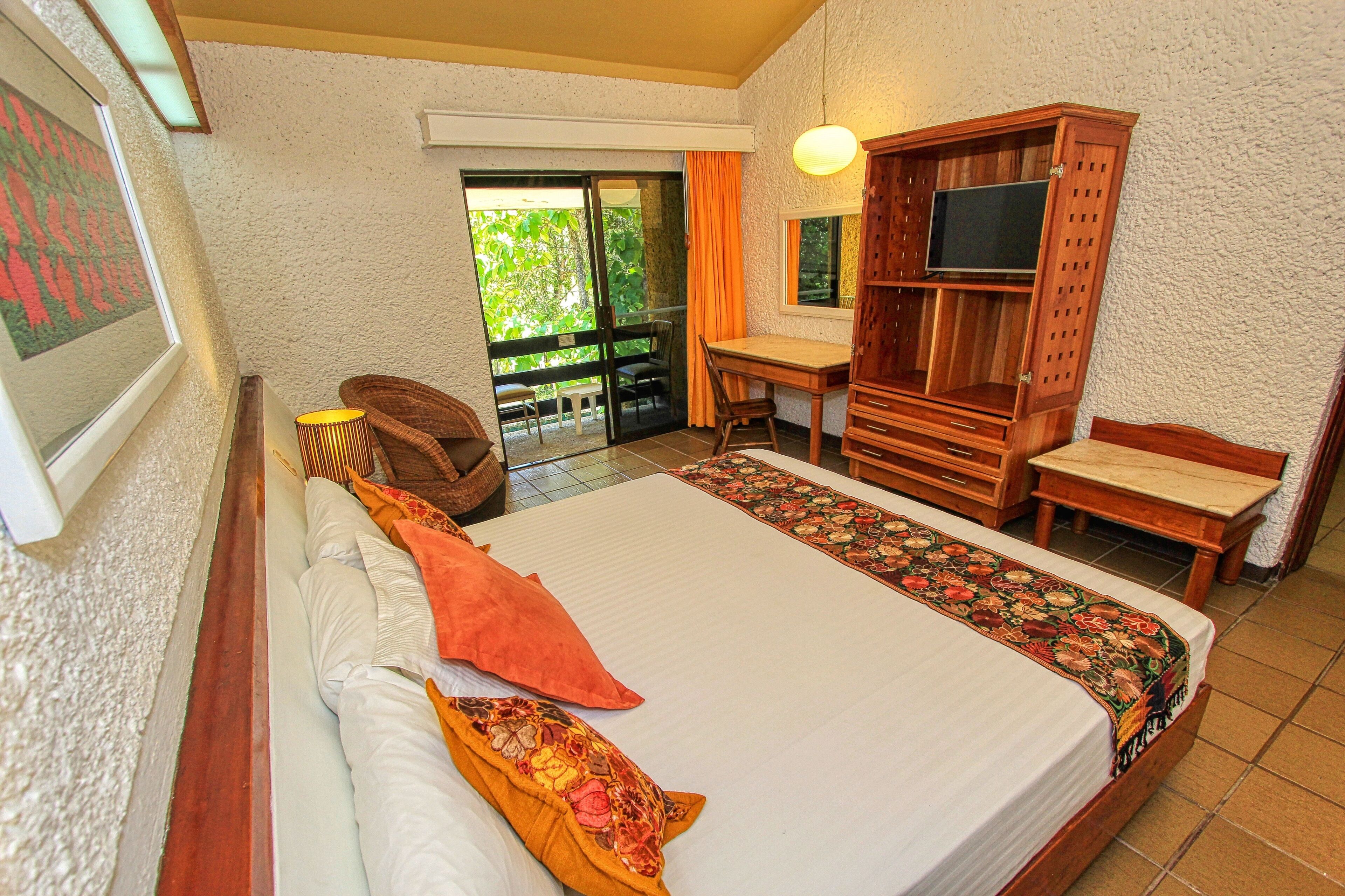 Habitación de hotel con cama, escritorio, televisor y balcón con vistas a la vegetación.