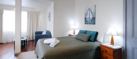 Habitación de hotel con cama, mesita de noche con lámpara, una silla y un cuadro en la pared.