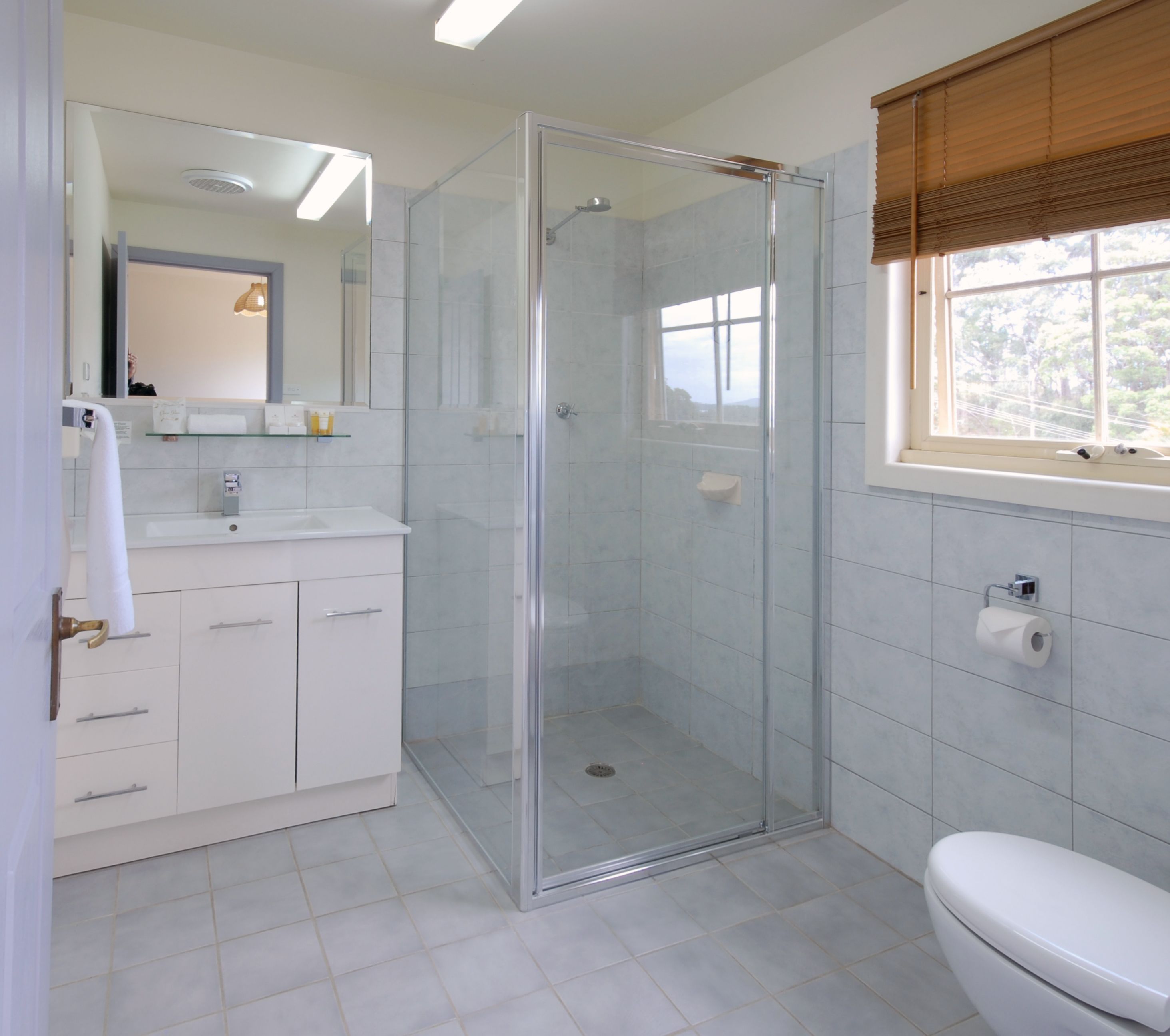 A bathroom with a glass-enclosed shower, a toilet, a sink with a mirror, and a window with blinds.