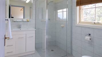 A bathroom with a glass-enclosed shower, a toilet, a sink with a mirror, and a window with blinds.