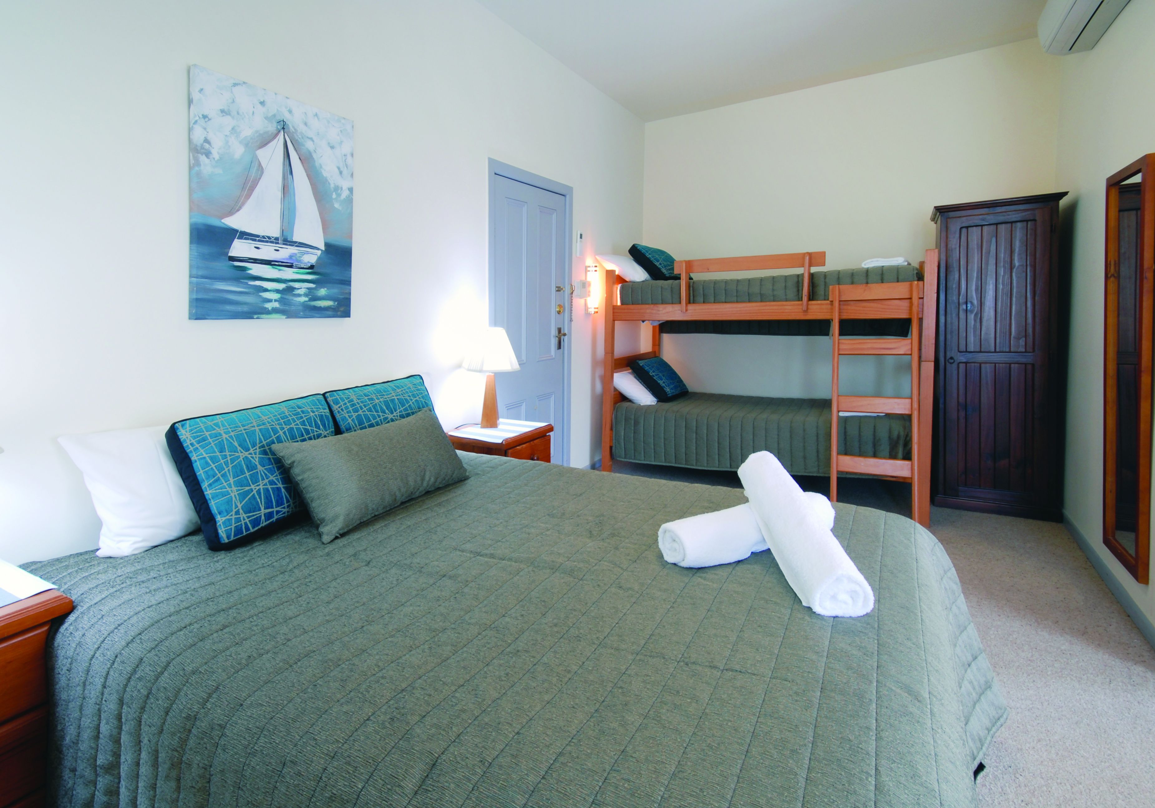 A room with a bunk bed, a single bed, a painting of a sailboat, a lamp, and a wardrobe.