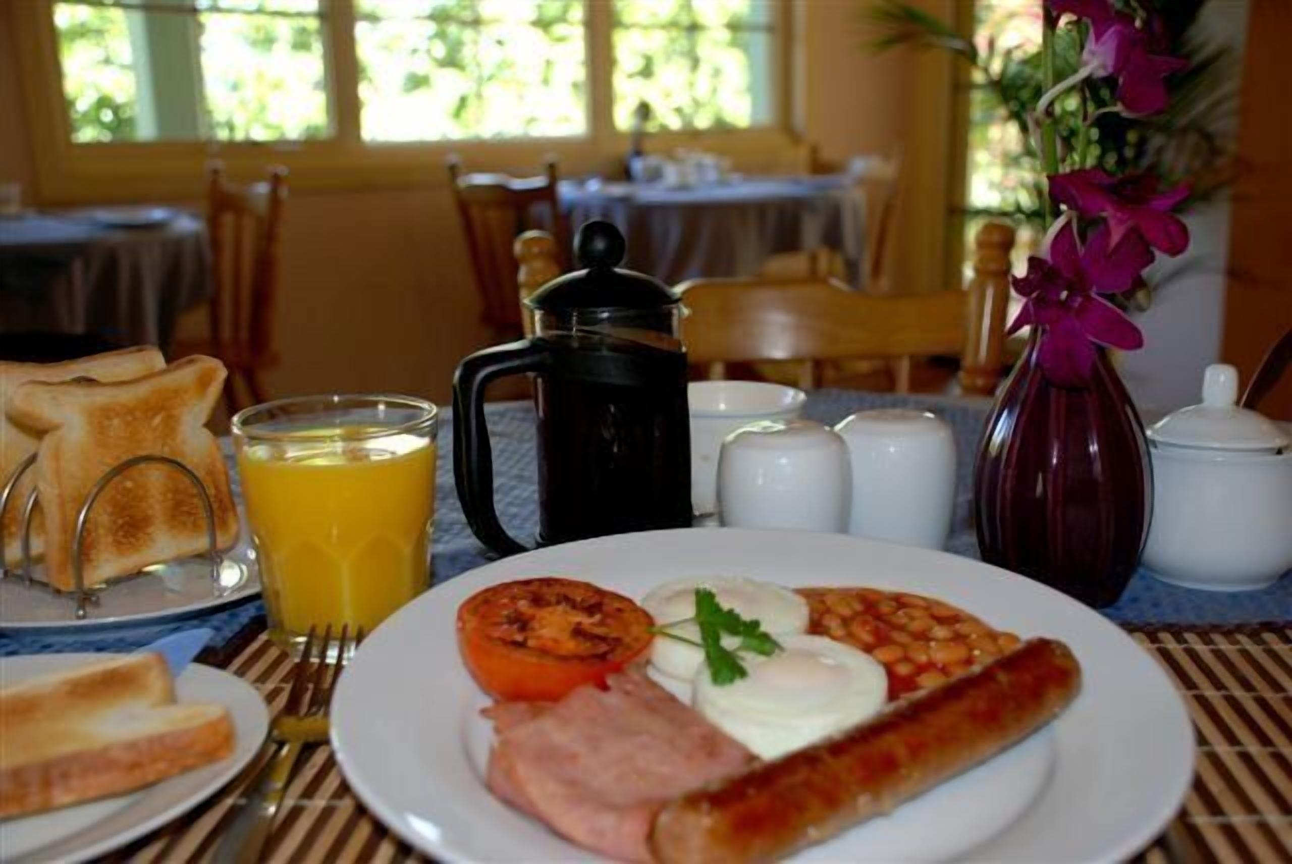 Ein Frühstückstisch mit Toast, Eiern, Schinken und einer Tomate auf einem Teller, ein Glas Orangensaft, eine Kaffeekanne und Salz- und Pfefferstreuer.