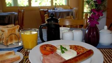 Colazione con pane tostato, uova, prosciutto e pomodoro su un piatto, un bicchiere di succo d'arancia, una caffettiera e salsiera e pepiera.