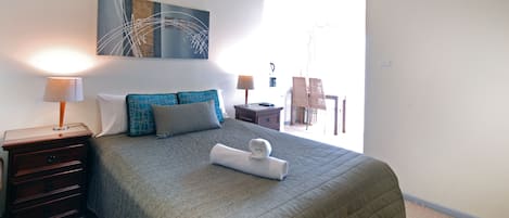 A bedroom with a bed, two bedside tables, a lamp, a chair, a painting, and a view of the outside.