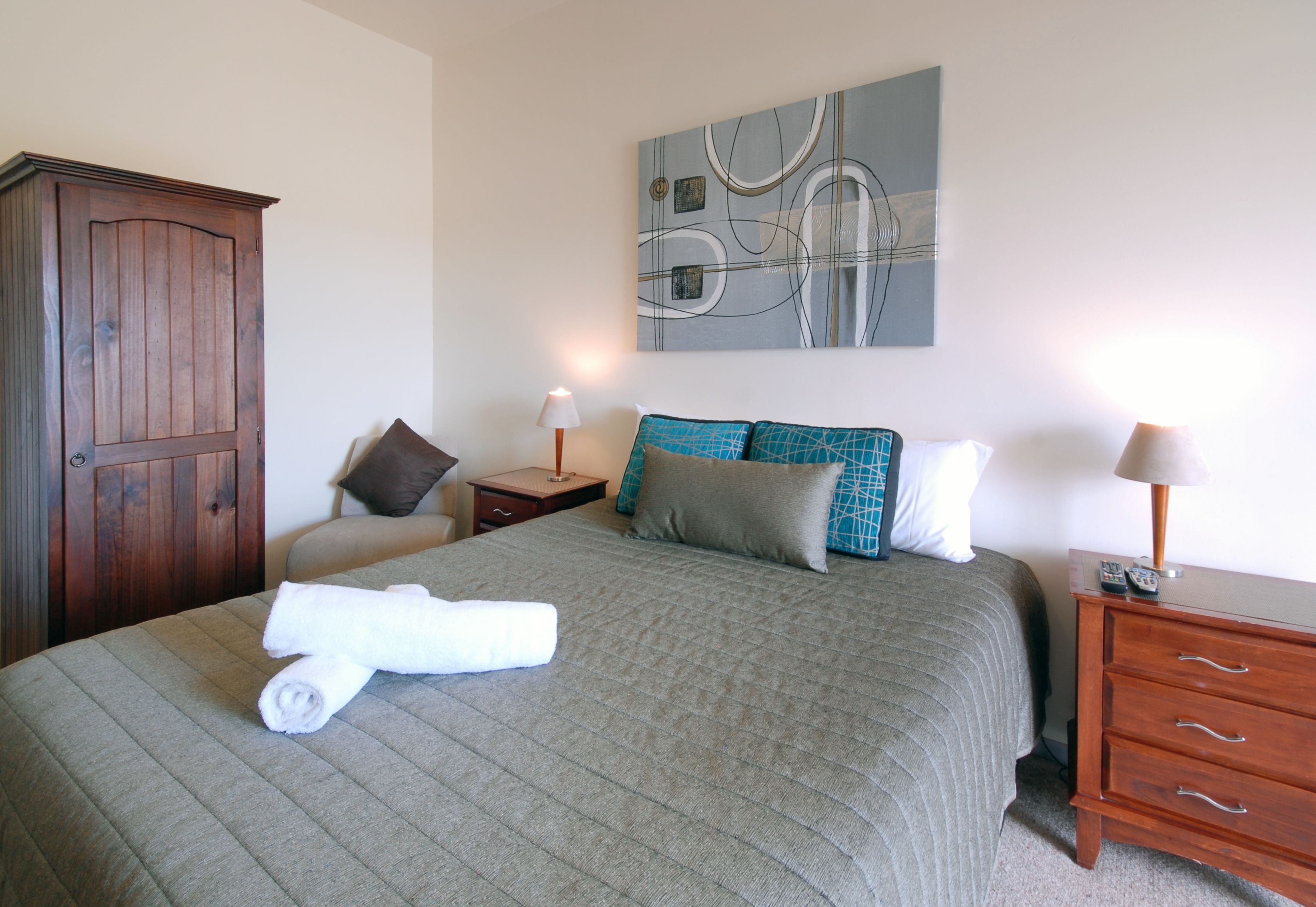 A neatly made bed with a grey quilt, a wooden wardrobe, a bedside table with a lamp, and a wall art piece.