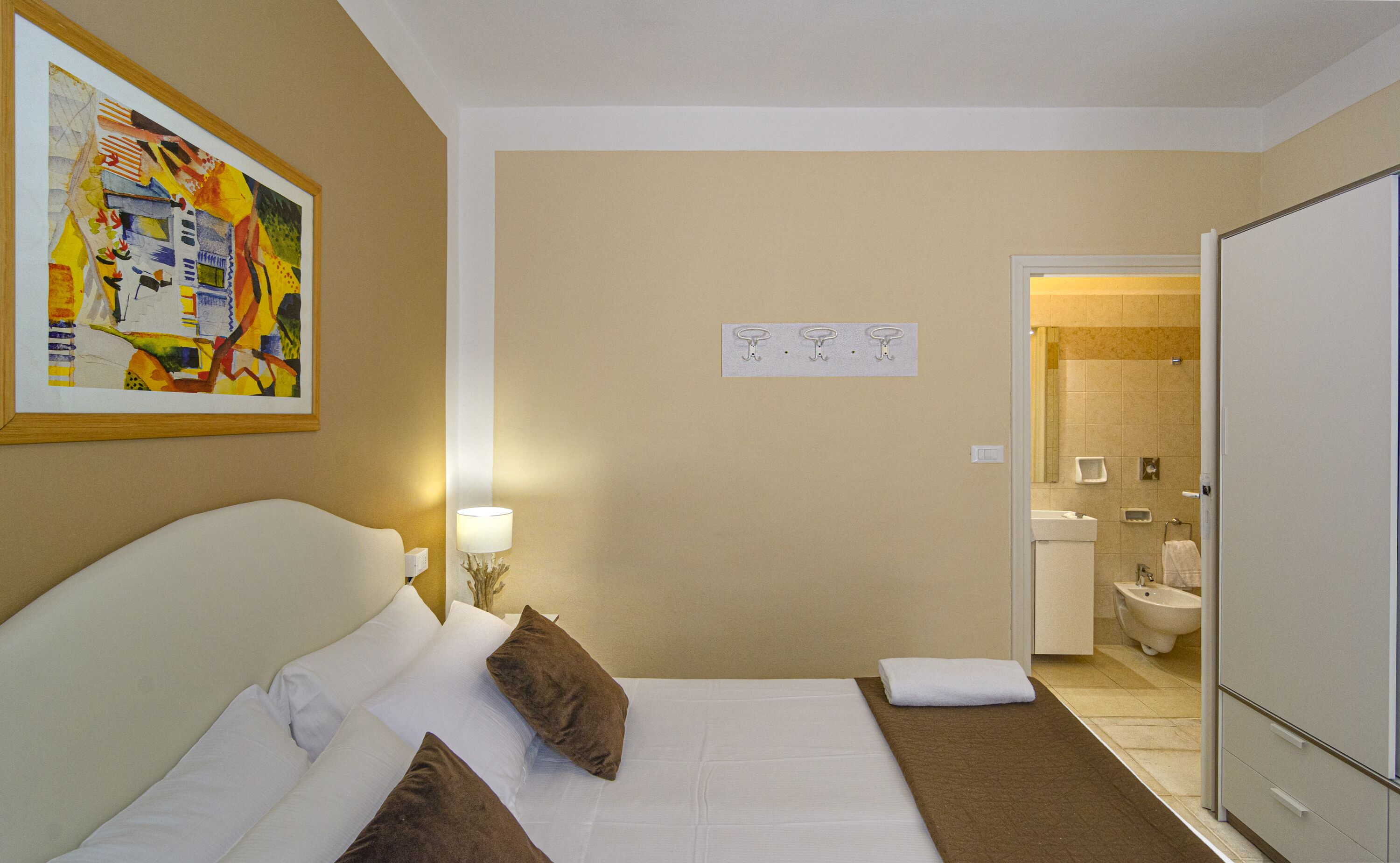 A hotel room with a bed, a painting on the wall, a lamp, and a bathroom visible through an open door.
