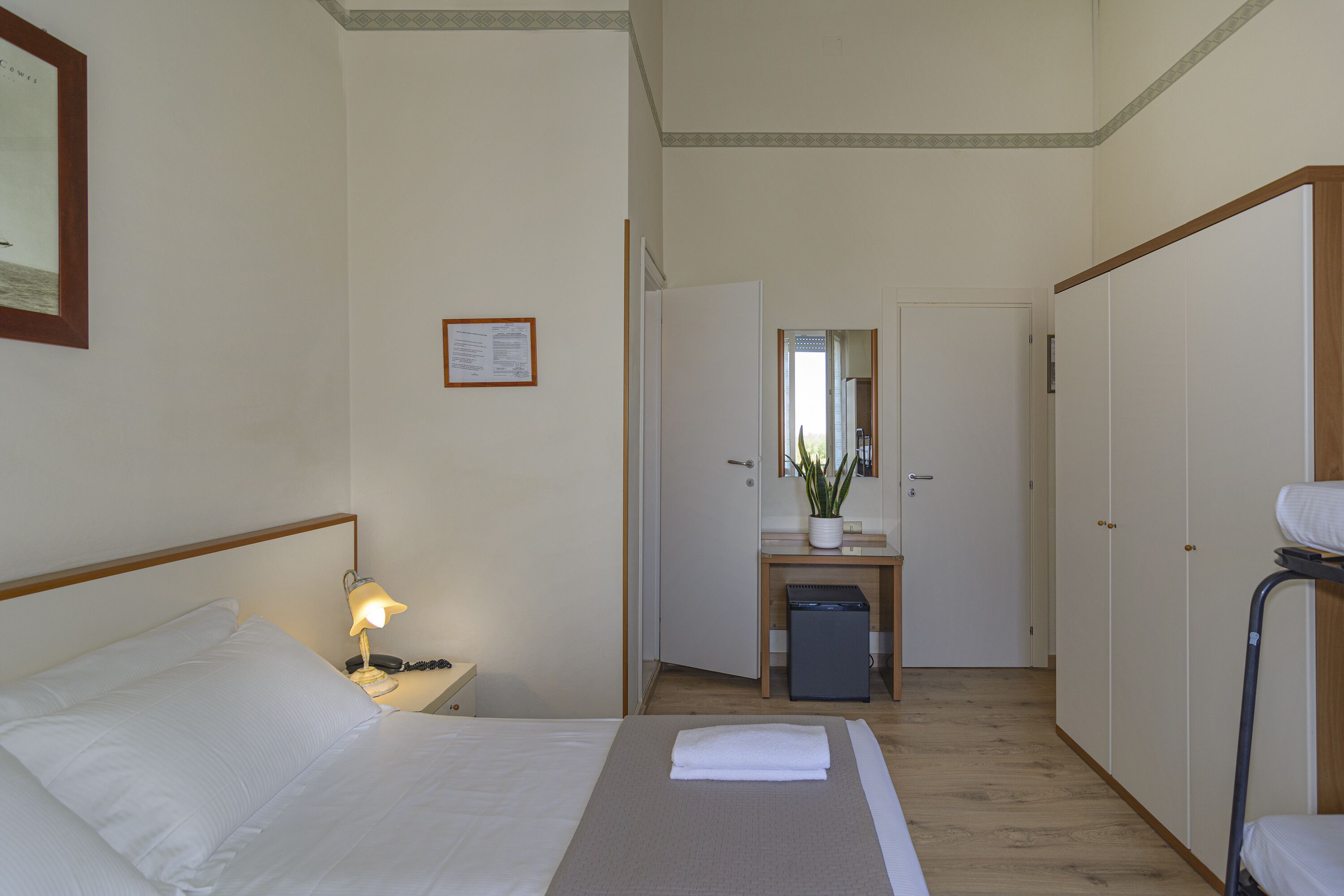 A hotel room with a bed, bedside table with a lamp, a desk with a mirror, a small refrigerator, and a wardrobe.