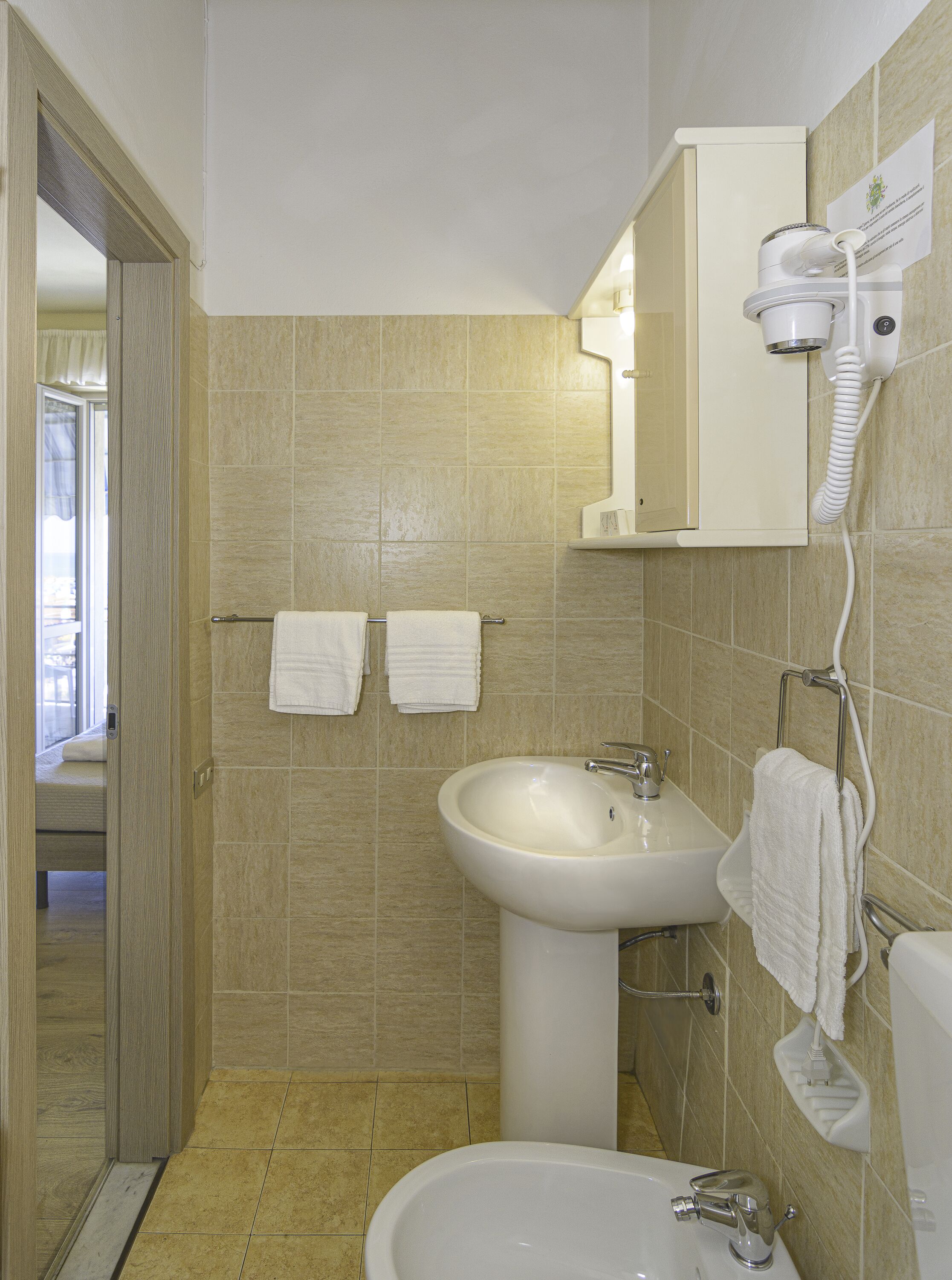 A bathroom with beige tiles, a sink, a mirror, a hairdryer, and two towels hanging on a rack.