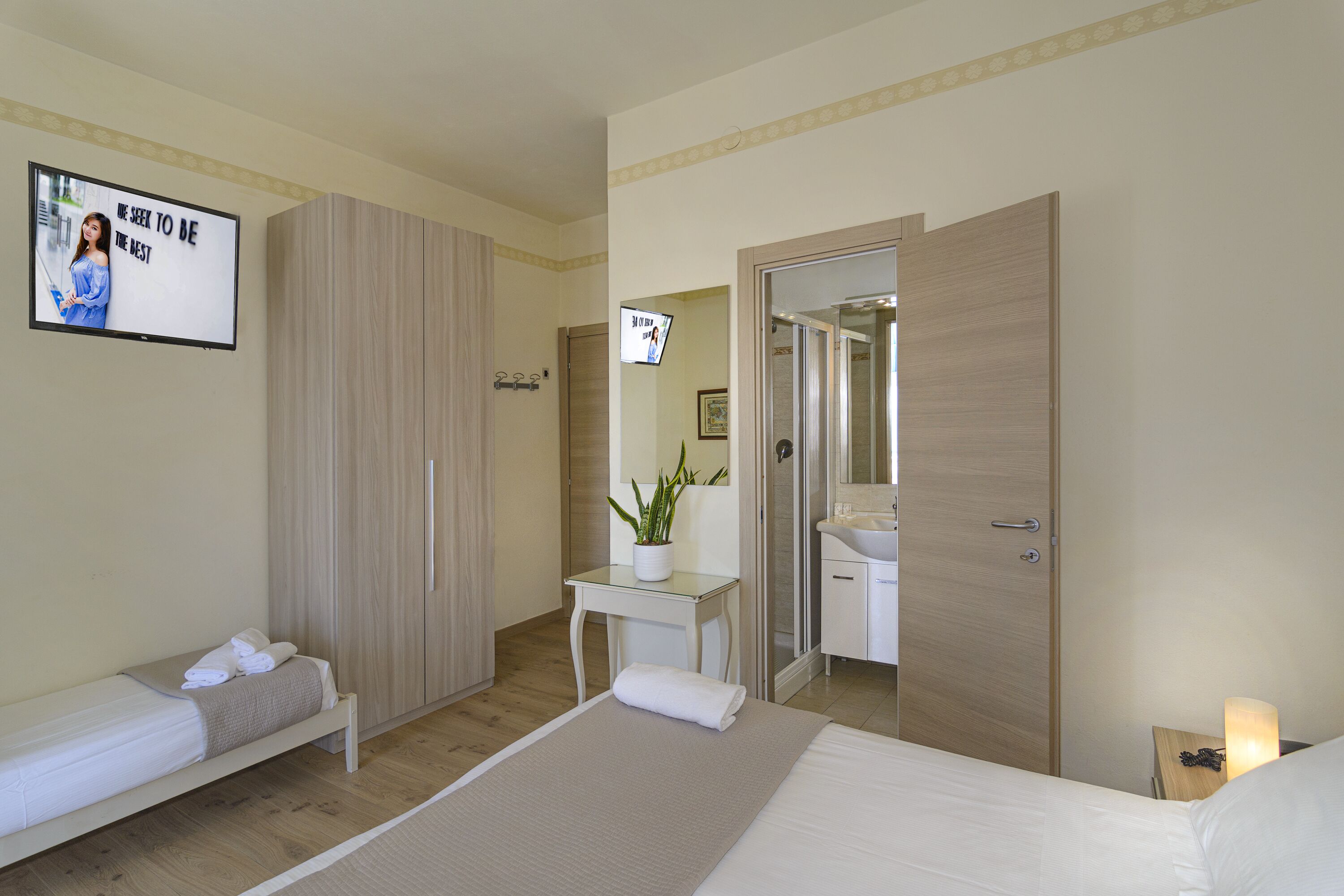A hotel room with a bed, a bedside table with a lamp, a small desk, a mirror, and a wardrobe.