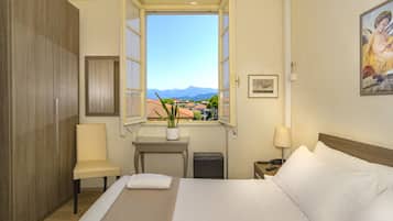 Quarto de hotel com cama, uma cadeira, uma mesinha com uma planta, janela com vista para montanhas e um quadro na parede.