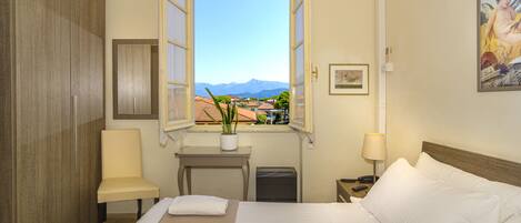 Una habitación de hotel con una cama, una silla, una mesita con una planta, una ventana que ofrece vistas a las montañas y un cuadro en la pared.