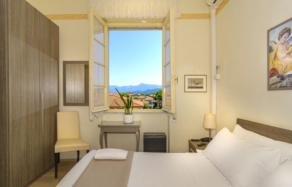 A hotel room with a bed, a chair, a small table with a plant, a window with a view of mountains, and a painting on the wall. - Hotel Lungomare (Camaiore)