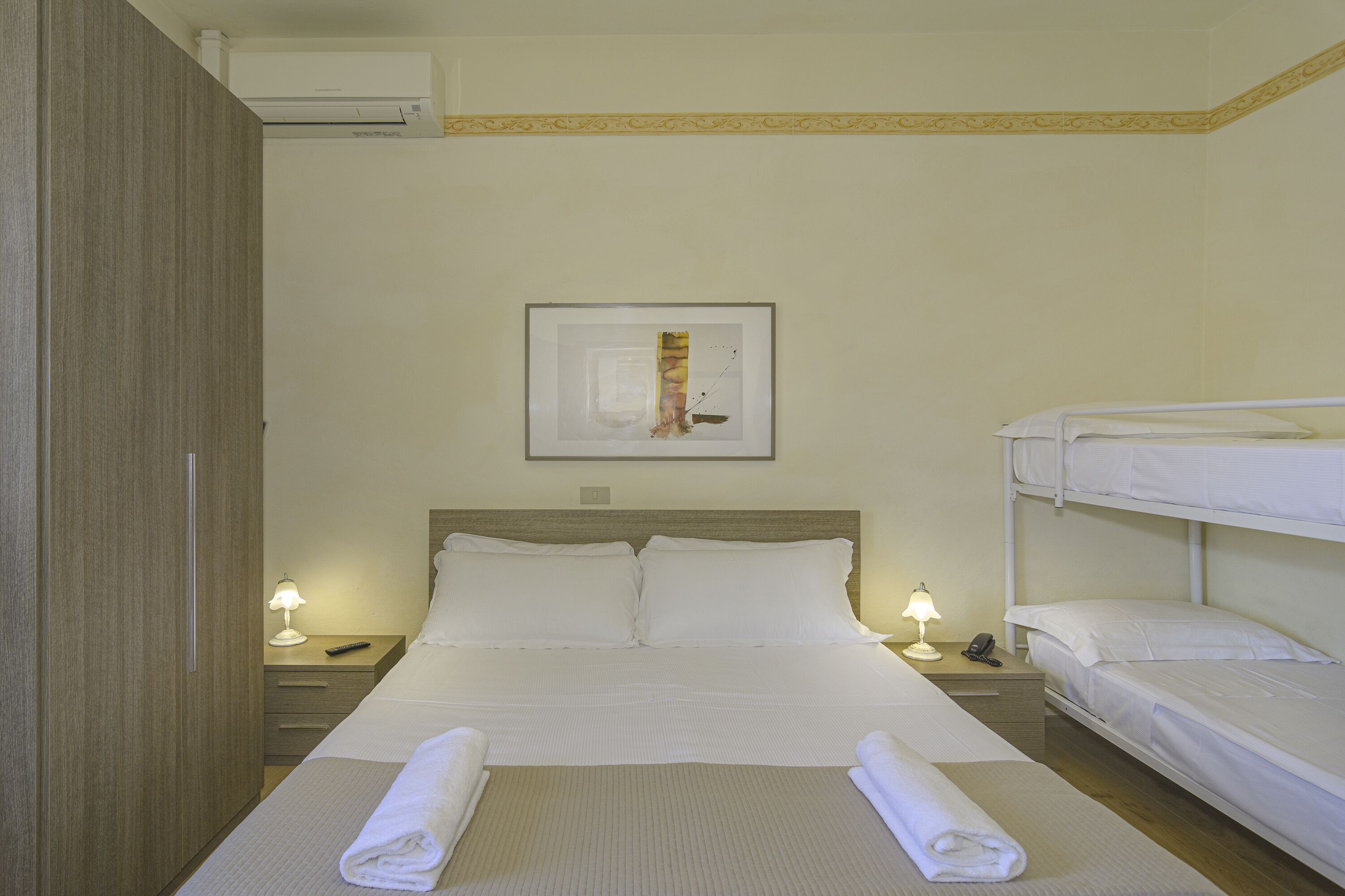 A hotel room with a single bed, a bunk bed, a bedside table with a lamp, and a framed picture on the wall.