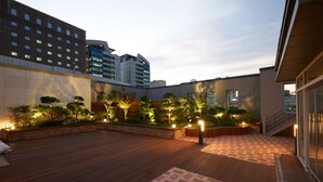 Terraza en la azotea con cubierta de madera, sendero de ladrillo y jardín ajardinado con árboles e iluminación.