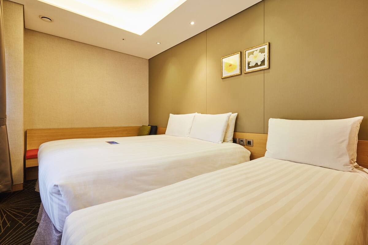 A hotel room with two beds, a wooden headboard, a ceiling light, and two framed pictures on the wall.