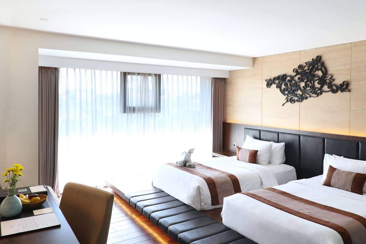 A hotel room with two beds, a large window with curtains, a desk with a vase of flowers, and a decorative wall piece.
