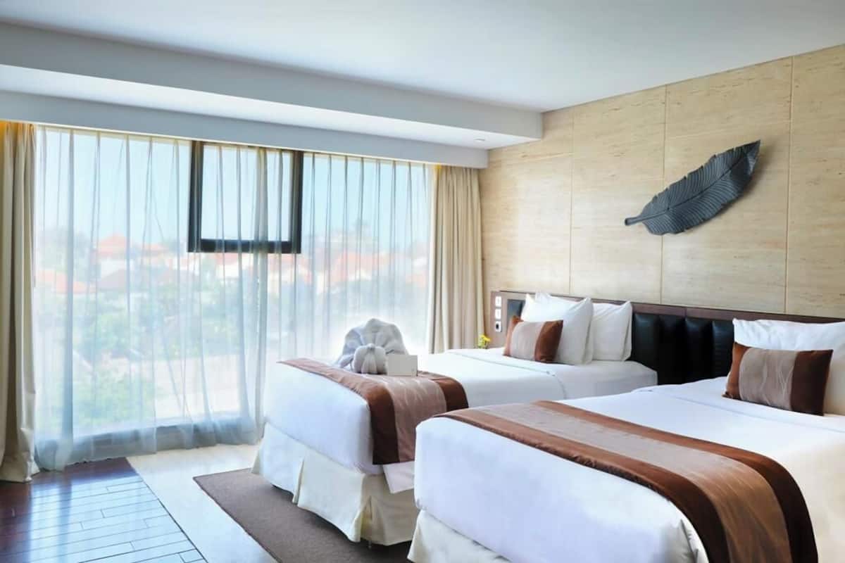 A hotel room with two beds, a large window with curtains, a wooden accent wall, and a leaf decoration.