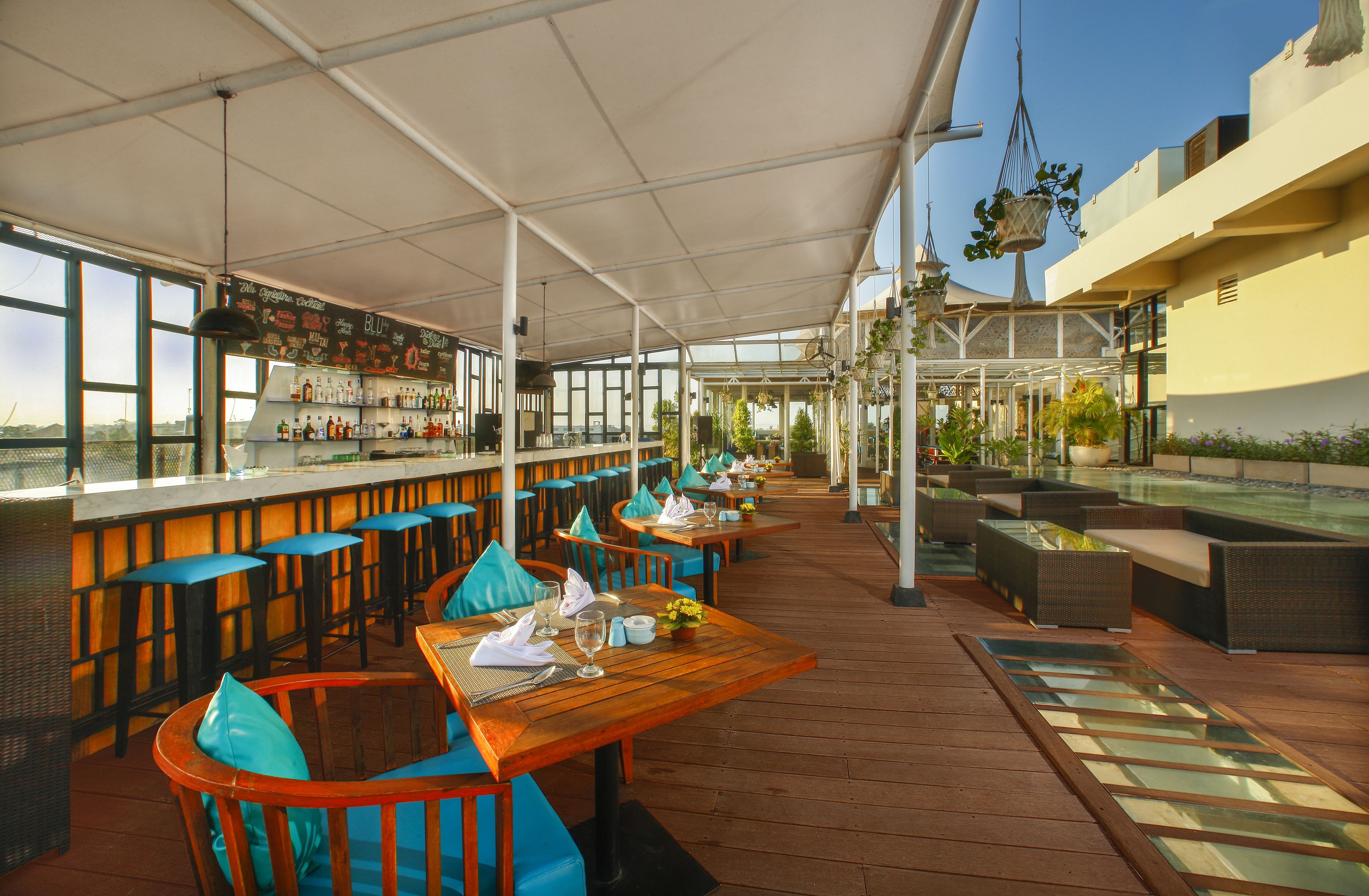An outdoor restaurant with wooden tables and blue cushioned chairs, a bar area, and a poolside seating area.