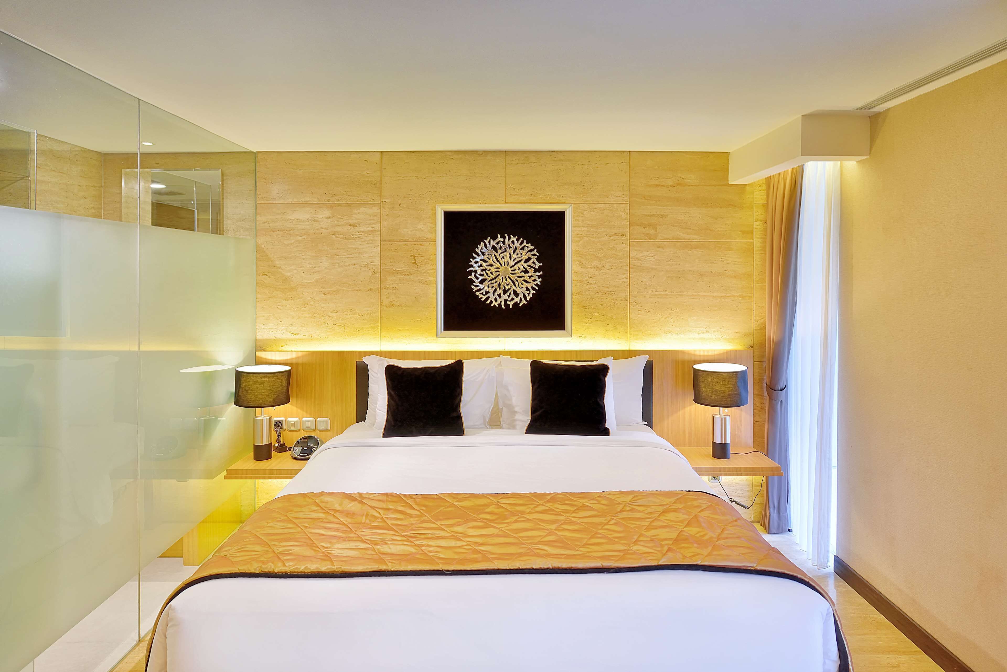 A hotel room with a large bed, a nightstand with a lamp, a wall-mounted artwork, and a glass partition.