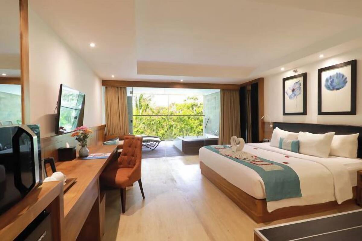 A hotel room with a bed, a desk with a TV, a chair, and a balcony with a view of greenery.