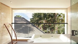 Une salle de bain moderne dotée d’une grande baignoire, d’une douche en verre et offrant une vue sur des palmiers et une maison, le tout visible à travers une grande fenêtre.