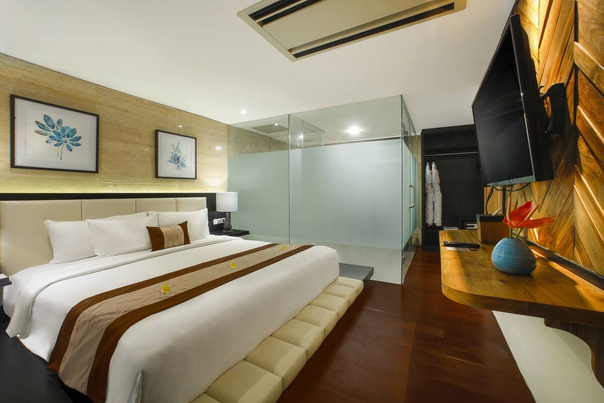 A modern hotel room with a large bed, a wooden headboard, a glass shower enclosure, a flat-screen TV mounted on the wall, and a wooden side table with a vase.