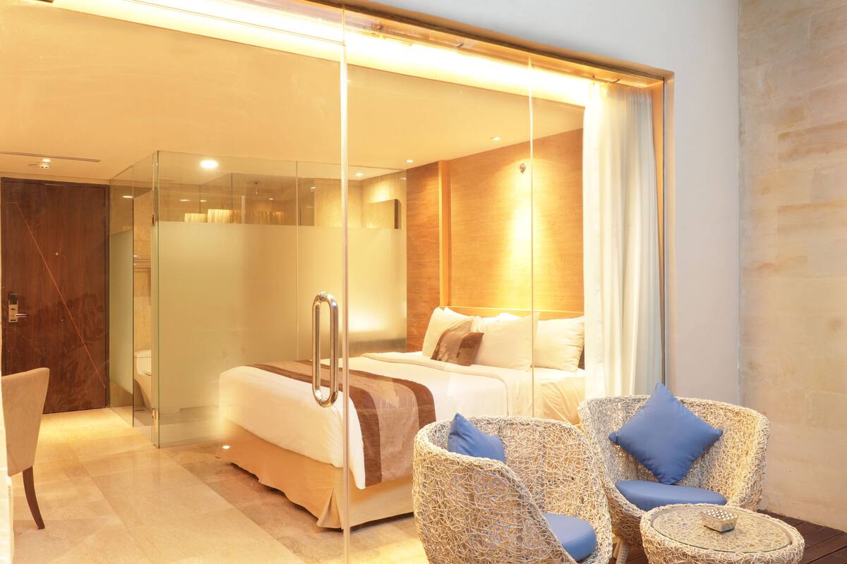 A modern hotel room with a glass partition, a bed, a seating area with wicker chairs, and a small round table.