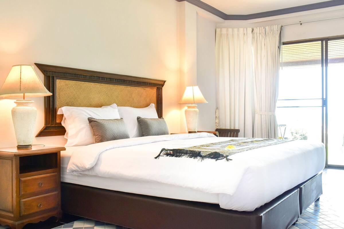 Ein Hotelzimmer mit einem großen Bett, zwei Nachttischlampen, einem hölzernen Kopfteil und Blick ins Freie durch ein Fenster mit Vorhängen.