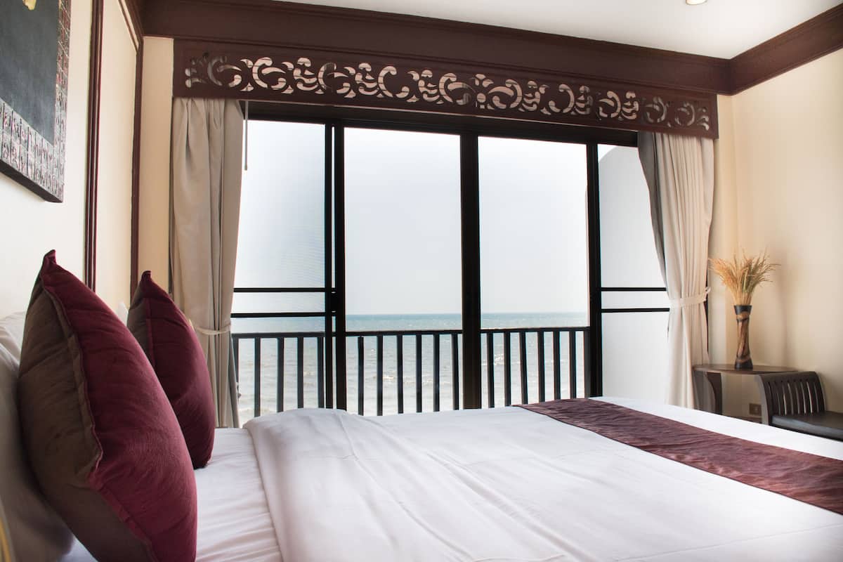 Ein Hotelzimmer mit einem großen Bett, zwei roten Kissen, Meerblick von einem Balkon und einem kleinen Tisch mit einer Vase.