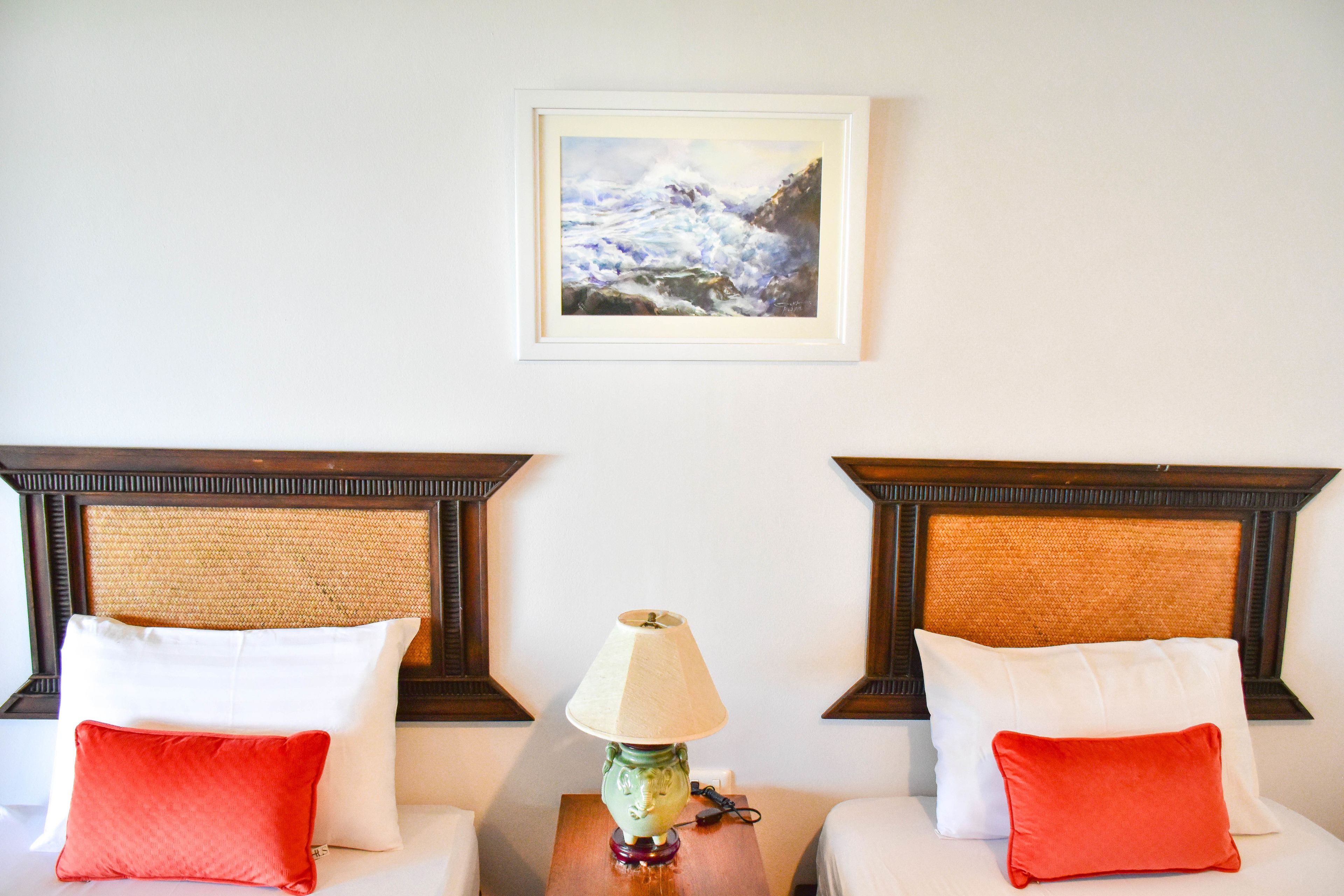 A hotel room with two beds, each with a white pillow and a red pillow, a small table with a lamp, and a framed painting of a stormy sea on the wall.