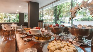 Disposición de un bufé con diversos postres y bebidas, sobre una mesa de madera en una habitación con ventanas amplias y sillas blancas.