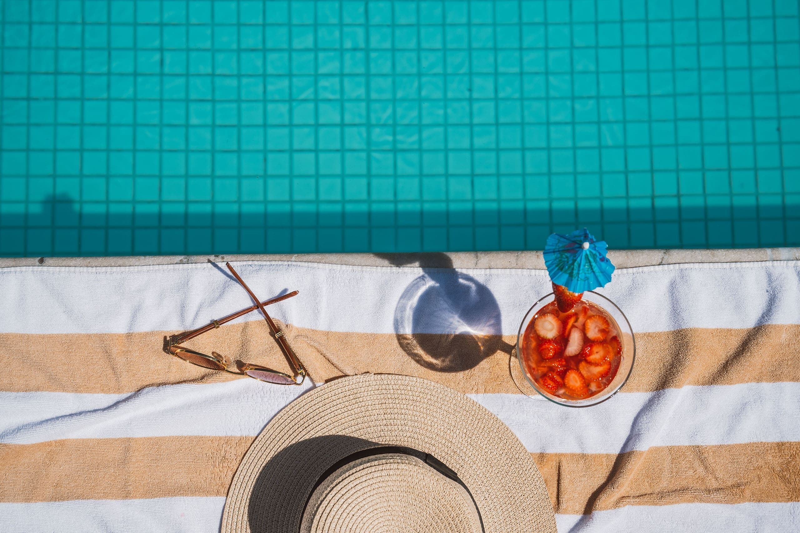 A striped beach towel, a straw hat, sunglasses, and a drink with a blue umbrella beside a pool.
