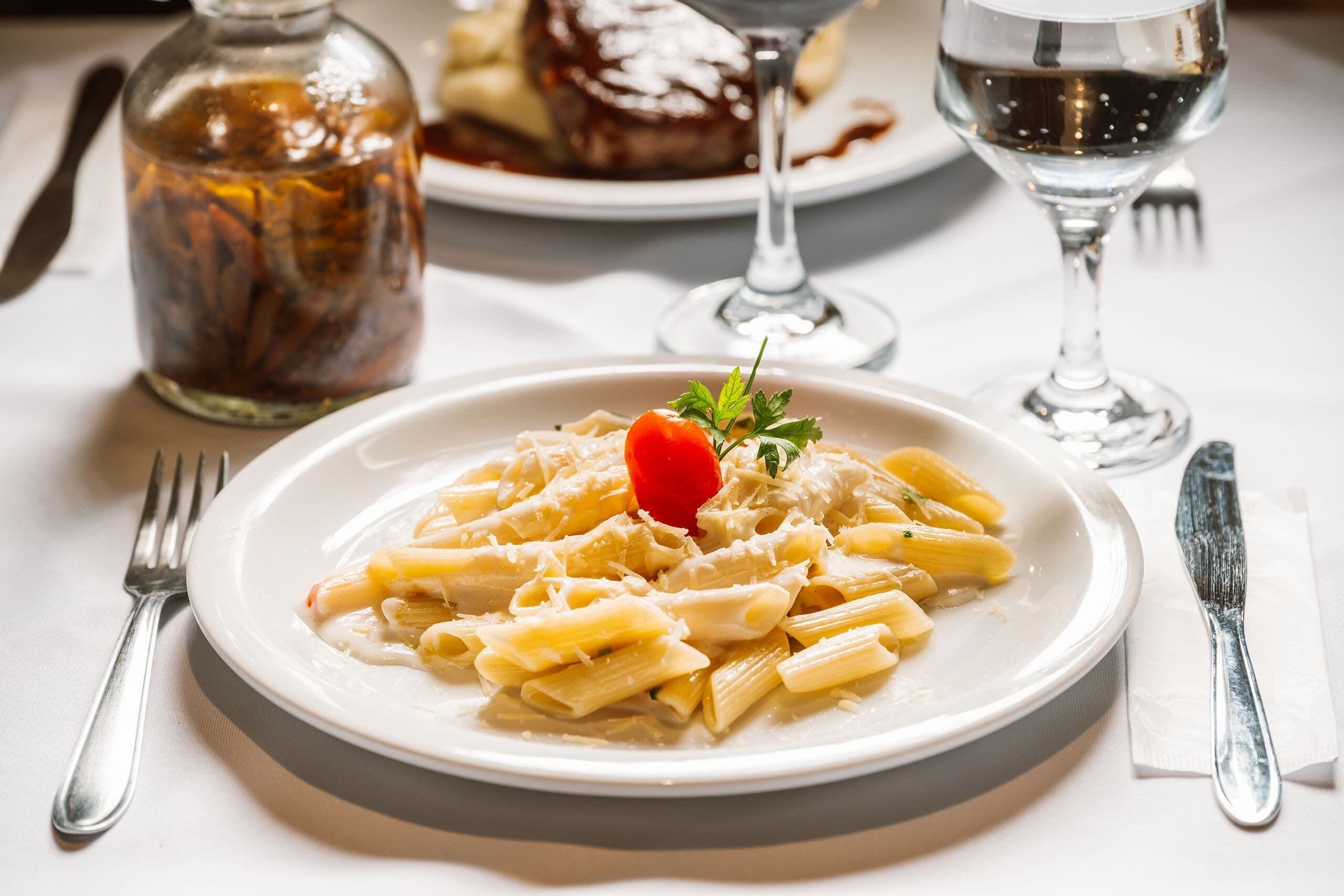 Une assiette de pâtes garnie de tomates cerises, un bocal de cornichons et un verre d’eau sur une table.