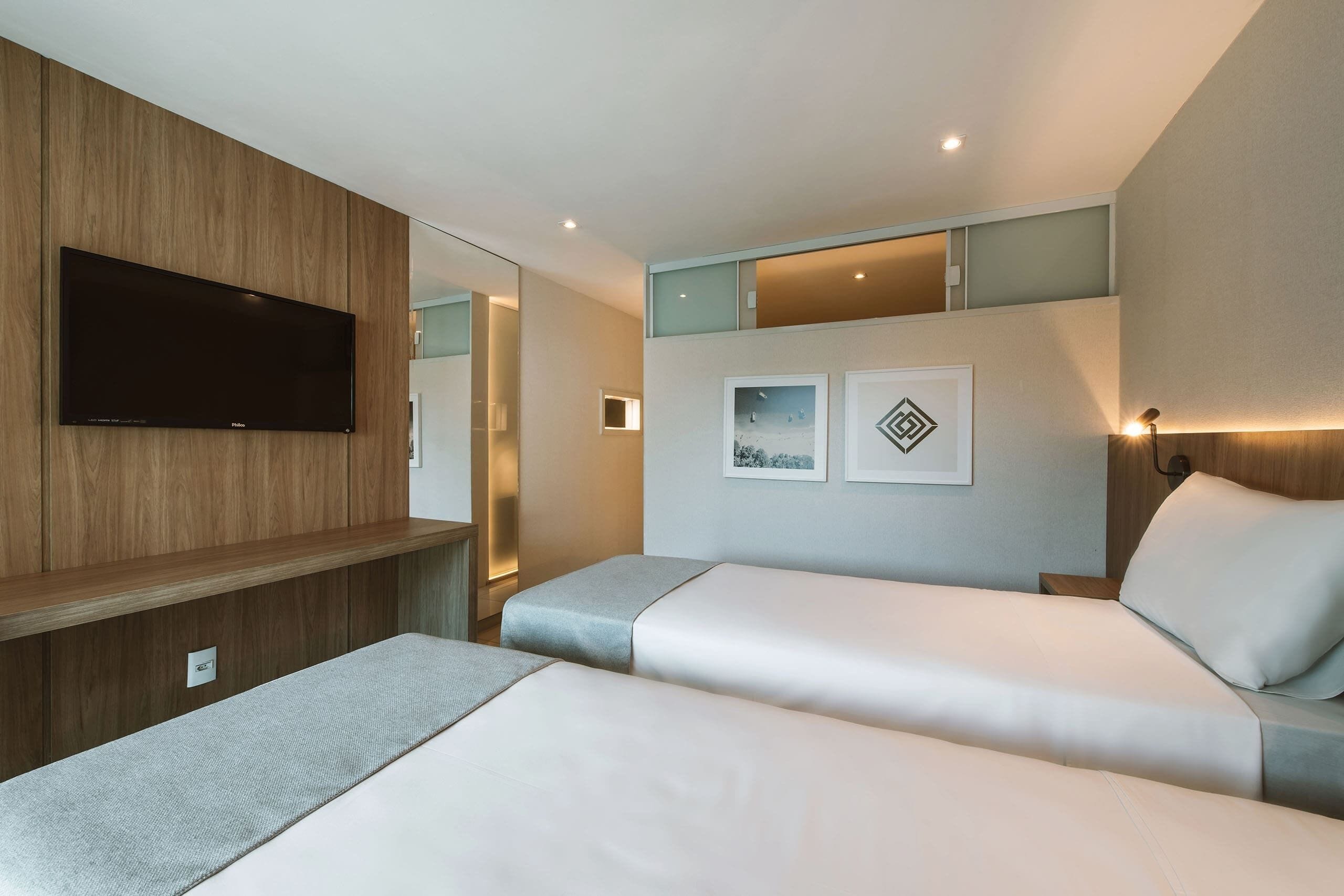 A hotel room with two beds, a mounted TV, and framed pictures on the wall.