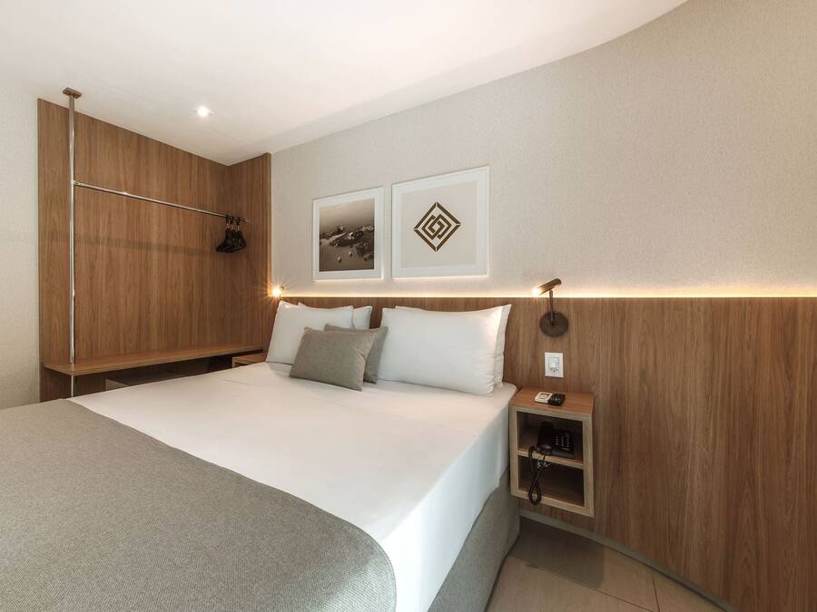 A modern hotel room with a large bed, a wooden wardrobe, a bedside table with a phone, and two framed pictures on the wall.