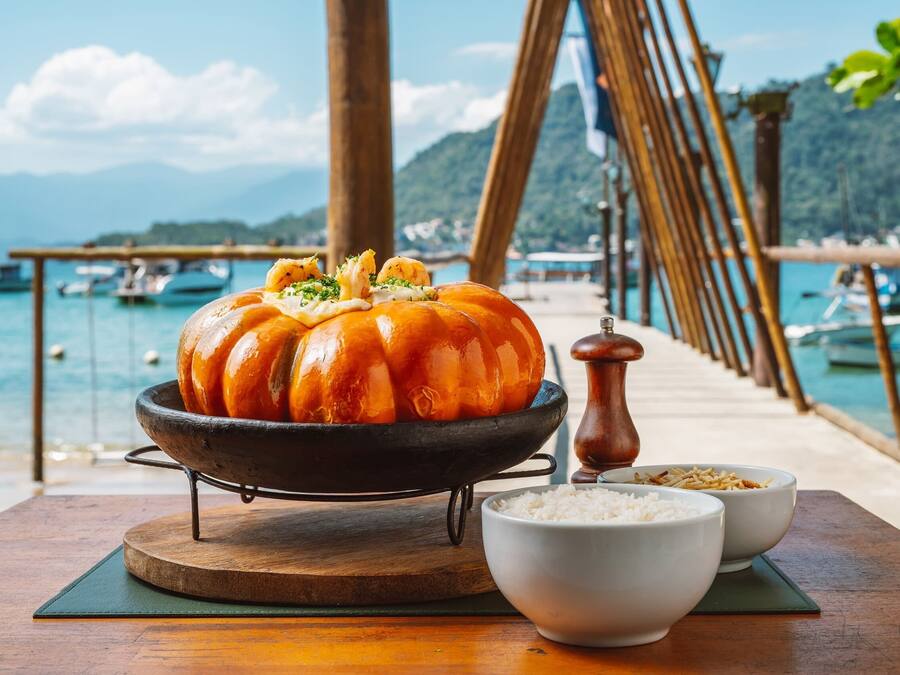 A pumpkin on a stand with bowls of rice and a pepper grinder on a wooden table by a waterfront.