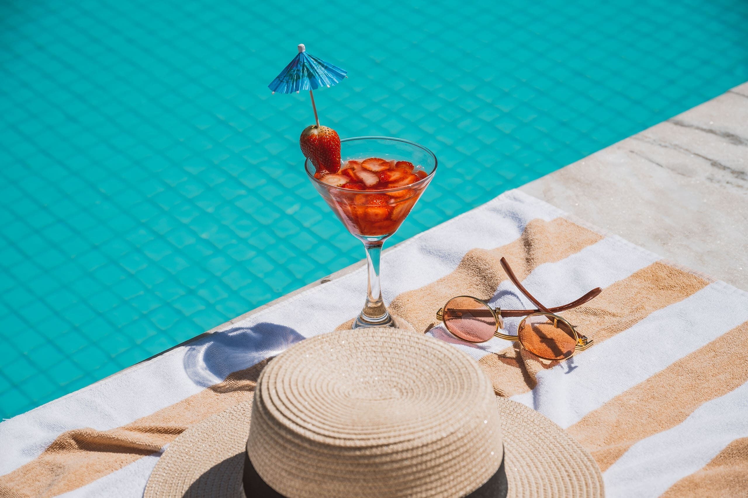 A cocktail with a strawberry garnish and a blue umbrella, sunglasses, and a straw hat on a striped towel by a pool.
