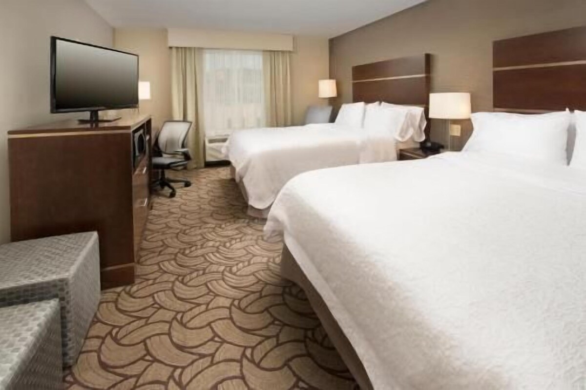 Quarto de hotel com duas camas, uma escrivaninha com cadeira, televisão e janela com cortinas.
